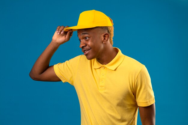 Delivery african american man in yellow polo shirt and cap looking aside touching his cap smiling friendly on isolated blue