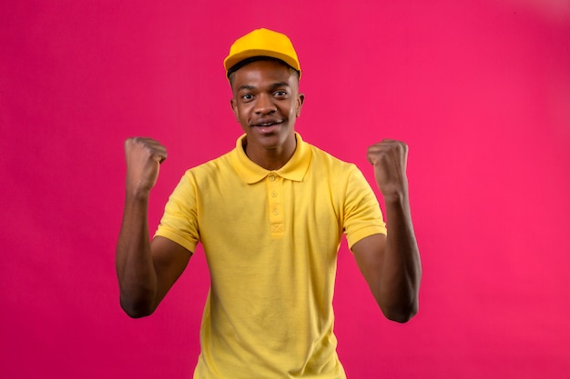 Афро-американский мужчина доставки в желтой рубашке поло и кепке выглядит взволнованным, радуясь своему успеху и победе, сжимая кулаки от радости, счастливым достичь своей цели и целей, стоя на розовом