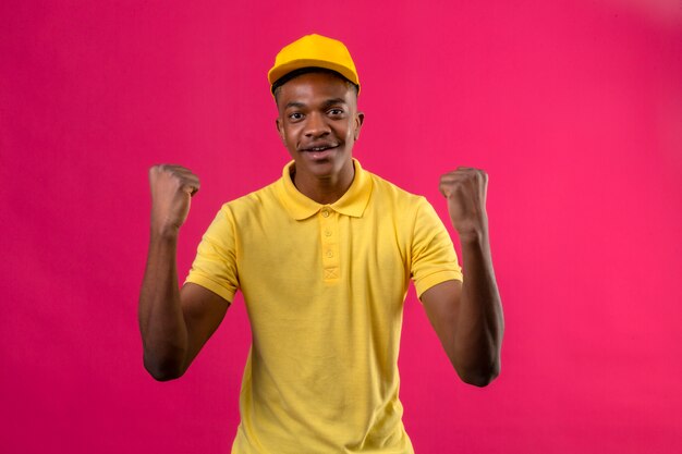 Delivery african american man in yellow polo shirt and cap looking exited rejoicing his success and victory clenching his fists with joy happy to achieve his aim and goals standing on pink