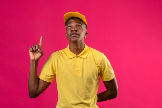 Delivery african american man in yellow polo shirt and cap looking up and pointing up with index finger standing on pink