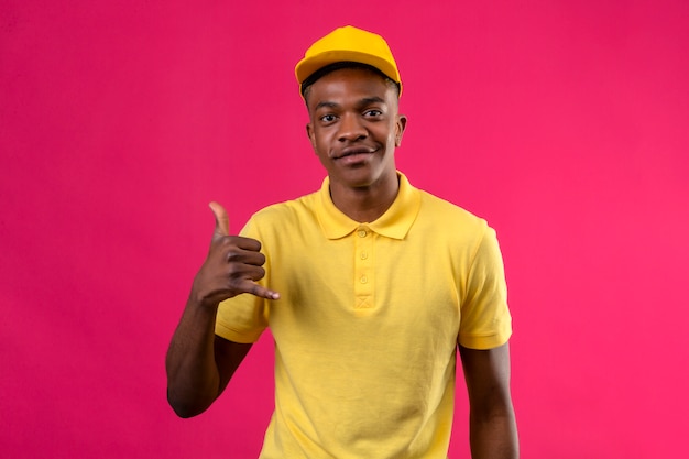 Delivery african american man in yellow polo shirt and cap making call me gesture looking confident smiling friendly on pink
