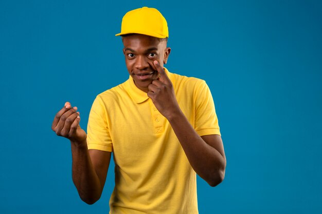 Delivery african american man in yellow polo shirt and cap making money gesture with hand pointing with finger to eye waiting for payment smiling on isolated blue