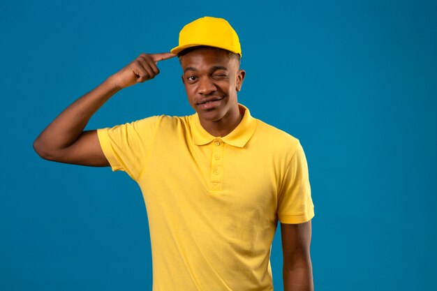 Афро-американский мужчина-доставщик в желтой рубашке-поло и кепке, указывающий на храм, подмигивает, радостно глядя на синее