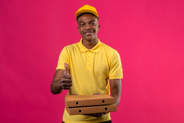 Delivery african american man in yellow polo shirt and cap smiling friendly holding pizza boxes showing thumbs up on pink