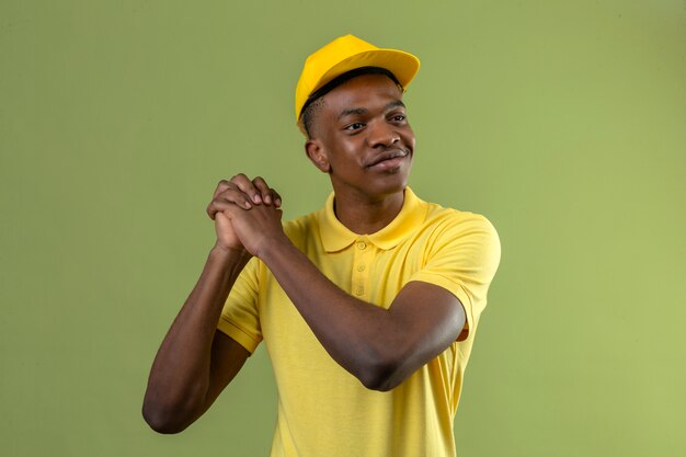 Delivery african american man in yellow polo shirt and cap smiling holding hands together grateful gesture standing on green