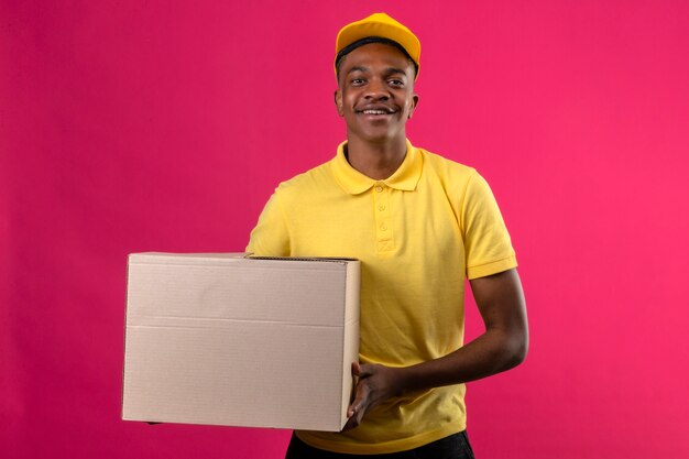 Delivery african american man in yellow polo shirt and cap standing with cardboard box smiling friendly on pink