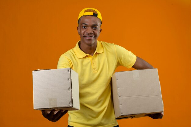 Афро-американский мужчина доставки в желтой рубашке поло и кепке, стоящий с картонными коробками в руках, дружелюбно улыбается на изолированном оранжевом