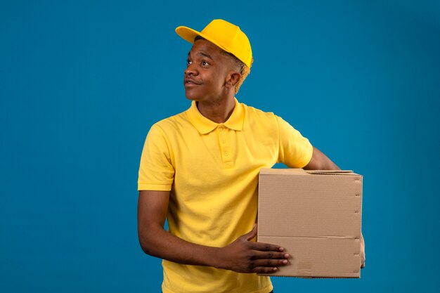 노란색 폴로 셔츠와 격리 된 파란색에 얼굴에 미소로 옆으로 찾고 골 판지 상자와 모자 서 배달 아프리카 계 미국인 남자