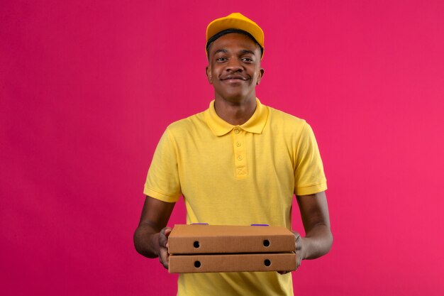 Delivery african american man in yellow polo shirt and cap standing with pizza boxes in hands smiling friendly on pink