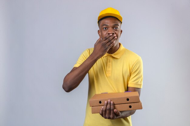 Delivery african american man in yellow polo shirt and cap standing with pizza boxes looking surprised coning mouth with hand standing