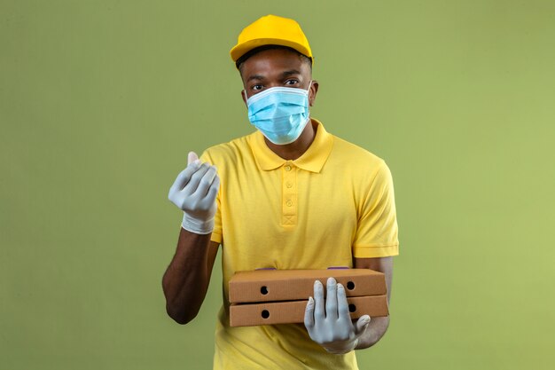 Delivery african american man in yellow polo shirt and cap wearing medical protective mask holding pizza boxes making money gesture with hand waiting for payment standing on green