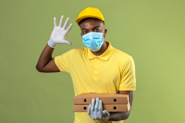 Delivery african american man in yellow polo shirt and cap wearing medical protective mask holding pizza boxes showing number five with open hand standing on green