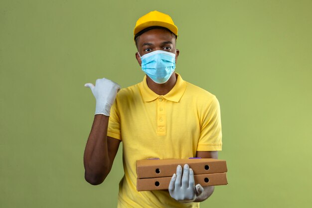 Delivery african american man in yellow polo shirt and cap wearing medical protective mask holding pizza boxes with serious face pointing to the side with thumb standing on green