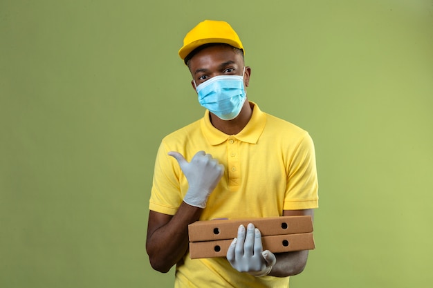 Афро-американский мужчина-доставщик в желтой рубашке поло и кепке в медицинской защитной маске держит коробки для пиццы с улыбкой на лице, указывая в сторону с большим пальцем, стоящим на зеленом