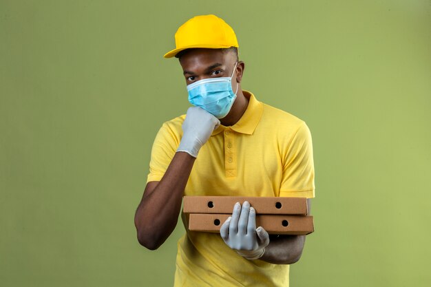 Delivery african american man in yellow polo shirt and cap wearing medical protective mask smiling and touching his chin on isolated green