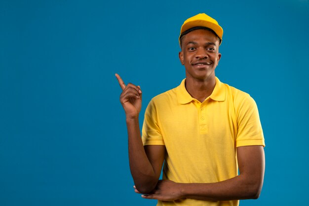 Афро-американский мужчина доставки в желтой рубашке поло и кепке с уверенной улыбкой, указывая пальцем в сторону, стоя на изолированном синем
