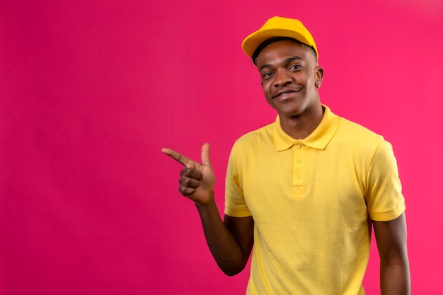 Delivery african american man in yellow polo shirt and cap with smile on face pointing with finger to the side standing on pink