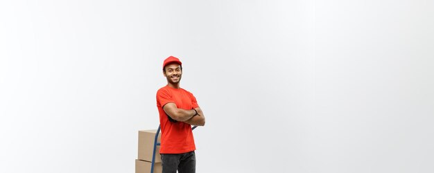 配達の概念ハンサムなアフリカ系アメリカ人の配達人または宅配便の肖像画は、灰色のスタジオの背景に分離されたボックスのスタックで手トラックを押す背景コピースペース