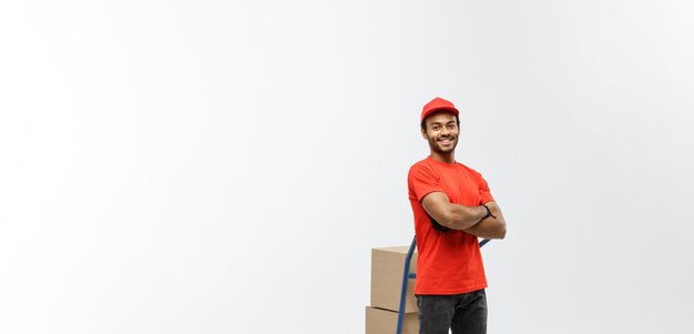 Delivery Concept Portrait of Handsome African American delivery man or courier pushing hand truck with stack of boxes Isolated on Grey studio Background Copy Space