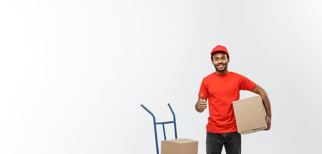 Free photo delivery concept portrait of handsome african american delivery man or courier with hand truck and holding box isolated on grey studio background copy space