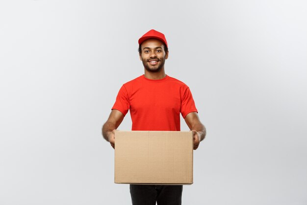 Концепция доставки - Портрет счастливый афро-американских человек доставки в синей ткани ходить, чтобы отправить пакет коробки для клиента. Изолированные на фоне серой студии. Копирование пространства.