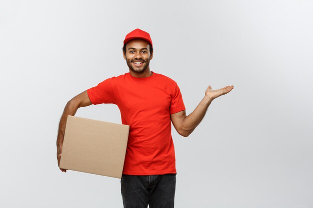 Концепция доставки - портрет счастливый афро-американских доставки человек, указывая руку, чтобы представить пакет коробки. Изолированные на фоне серой студии. Копирование пространства.