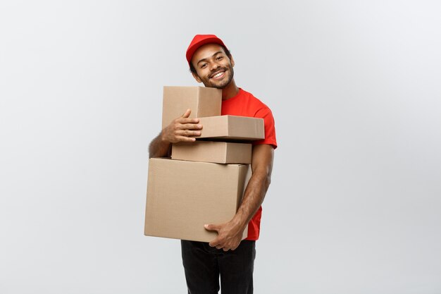 配達の概念 - ボックスパッケージを保持している赤い布でアフリカ系アメリカ人の配達人の肖像画の肖像画。灰色のスタジオの背景に分離。スペースをコピーします。