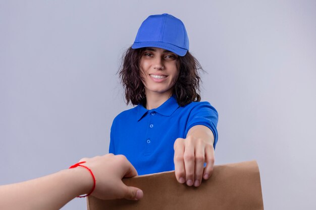 Ragazza delle consegne in uniforme blu e cappuccio che dà il pacchetto di carta a un cliente sorridente amichevole su bianco isolato