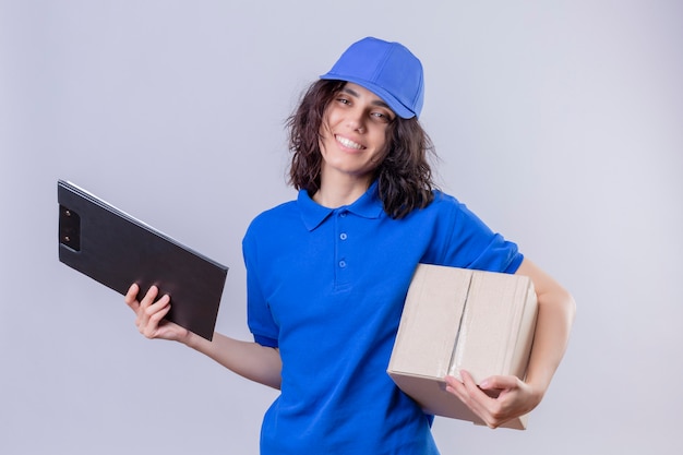 Ragazza di consegna in uniforme blu e pacchetto della scatola della tenuta del cappuccio e in piedi amichevole sorridente della lavagna per appunti su bianco