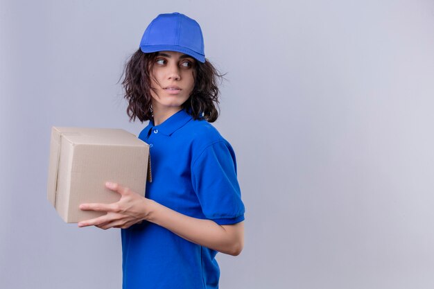 Delivery girl in blue uniform and cap holding box package standing sideways looking away with serious face on white