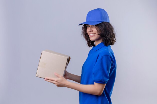 Delivery girl in blue uniform and cap holding box package standing sideways looking confident on isolated white