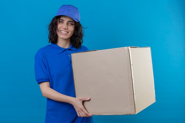 Доставщица в синей форме и кепке держит большую картонную коробку с улыбкой на лице, позитивно и счастливо стоя на синем