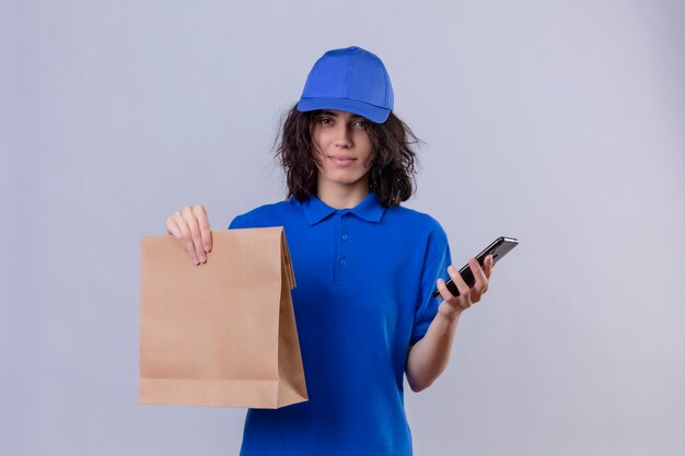 Delivery girl in blue uniform and cap holding paper package and mobile phone  with serious confident expression standing 