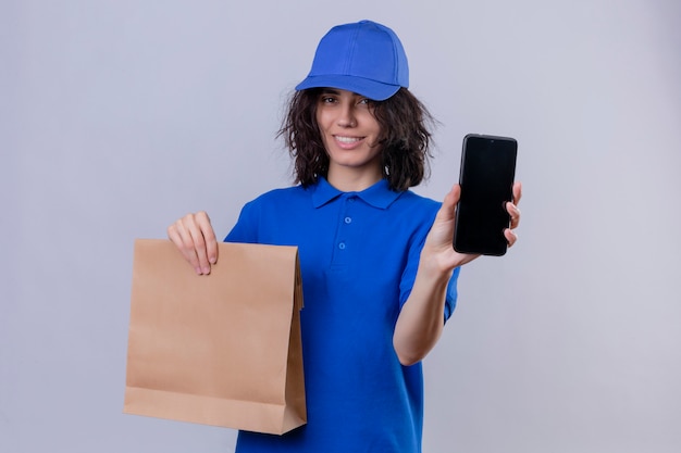 Ragazza di consegna in uniforme blu e cappuccio che tiene il pacchetto di carta che mostra il telefono cellulare che sorride allegramente in piedi sul bianco