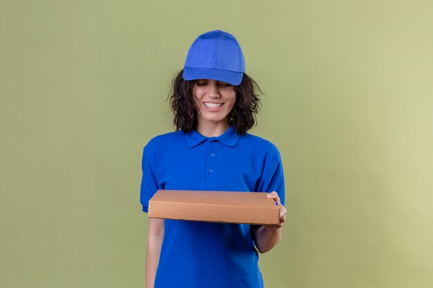 Delivery girl in blue uniform and cap holding pizza box smiling cheerful looking at box standing on isolated olive color