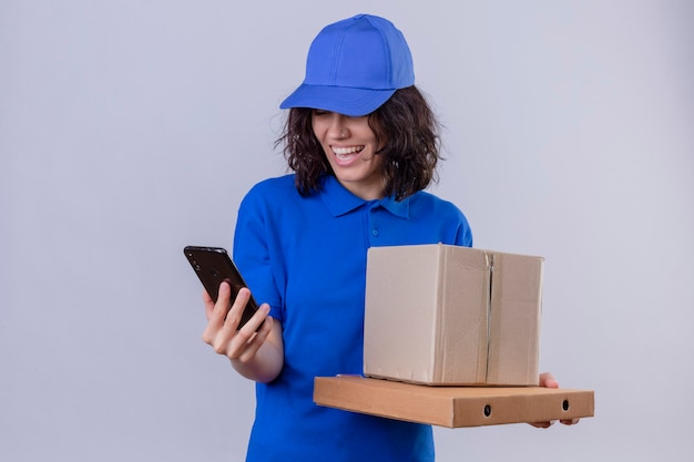 Delivery girl in blue uniform and cap holding pizza boxes and box package looking at screen of mobile phone smiling with happy face standing 