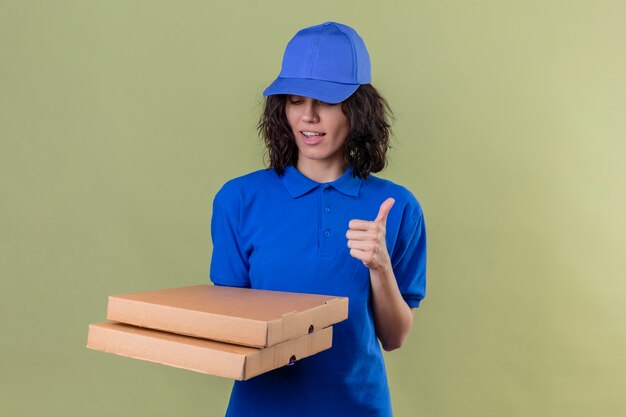 Delivery girl in blue uniform and cap holding pizza boxes looking down showing thumbs up smiling confident standing over olive color space