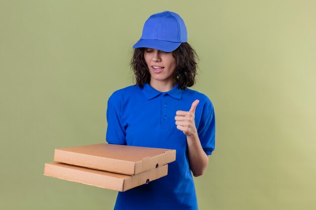 Доставщица в синей форме и кепке держит коробки для пиццы, глядя вниз, показывая пальцы вверх, улыбаясь уверенно, стоя на оливковом цвете