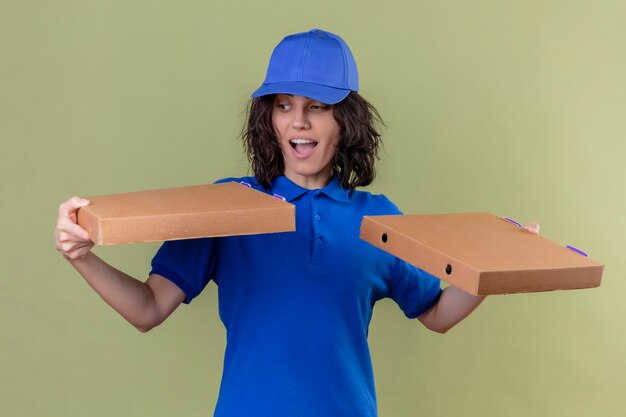 青い制服を着た配達少女と探しているピザの箱を保持しているキャップが終了し、孤立したオリーブ色のスペースの上に立って驚いた