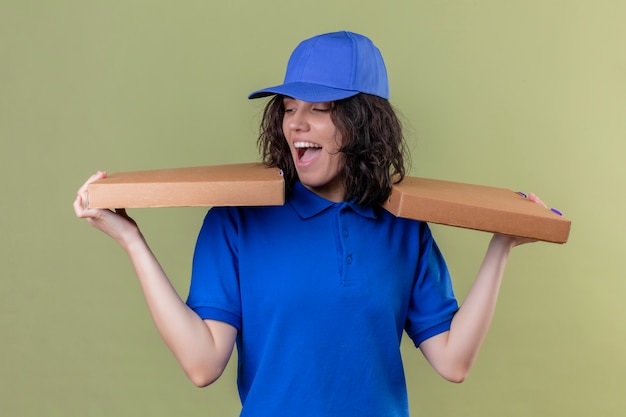 Ragazza delle consegne in uniforme blu e cappuccio che tiene le scatole per pizza che sembrano gioiosa sorridente positiva e felice allegramente in piedi sul verde isolato