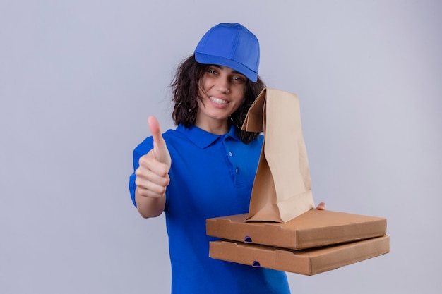 Доставщица в синей форме и кепке держит коробки для пиццы и бумажный пакет с улыбкой на лице, показывая большие пальцы руки вверх, стоя на белом