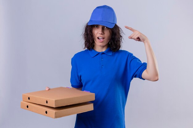 青い制服と立っているアイデアに集中して指で寺院を指しているピザの箱を保持しているキャップの配達の少女
