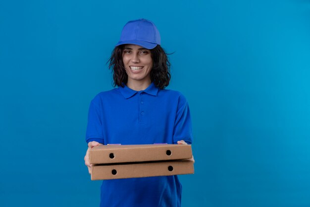 Ragazza delle consegne in uniforme blu e cappuccio che tiene le scatole per pizza in piedi amichevole sorridente positivo e felice sopra lo spazio blu isolato