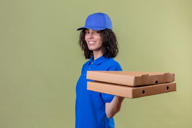Доставщица в синей форме и кепке держит коробки для пиццы позитивно и счастливо улыбается, дружелюбно стоит над изолированной зеленой зоной