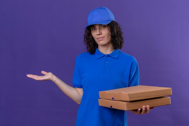 Delivery girl in blue uniform and cap holding pizza boxes and presenting with arm of the hand smiling friendly standing over purple space
