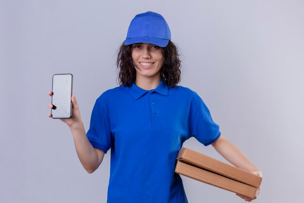 Delivery girl in blue uniform and cap holding pizza boxes showing mobile phone smiling friendly standing on white