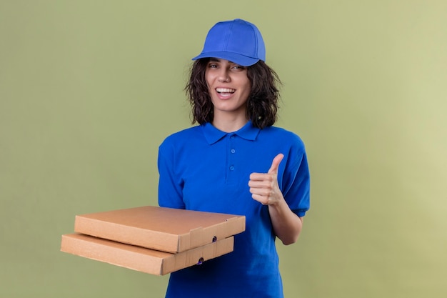 Доставщица в синей форме и кепке держит коробки для пиццы и весело улыбается, показывая большие пальцы руки вверх, стоя над изолированной зеленой зоной