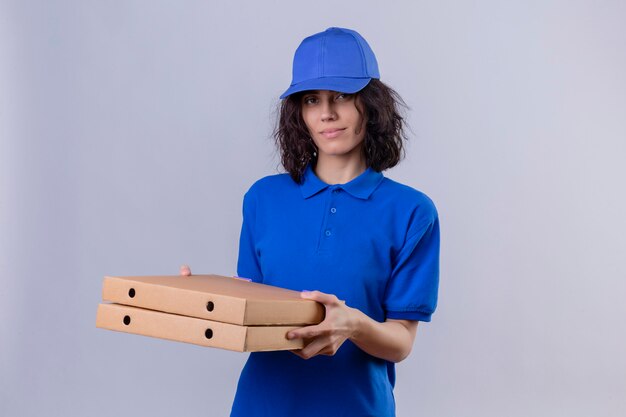 Доставщик в синей форме и кепке держит коробки для пиццы, уверенно улыбаясь, стоя на белом