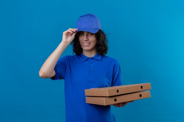 Delivery girl in blue uniform and cap holding pizza boxes with friendly smile touching her cap standing on isolated blue