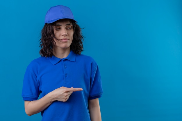 Foto gratuita ragazza delle consegne in uniforme blu e berretto che osserva da parte con espressione sospettosa che punta con il dito indice di lato in piedi sul blu